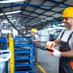 Factory worker wearing uniform and hardhat operating industrial machine with push button joystick in production hall.